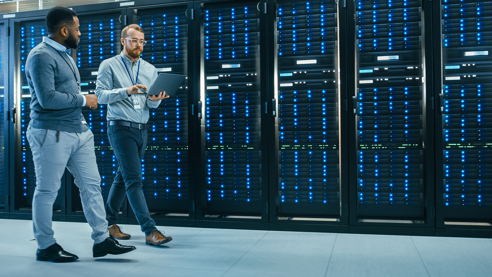 Two men standing in a data center, positioned in front of rows of servers, engaged in discussion or monitoring equipment.