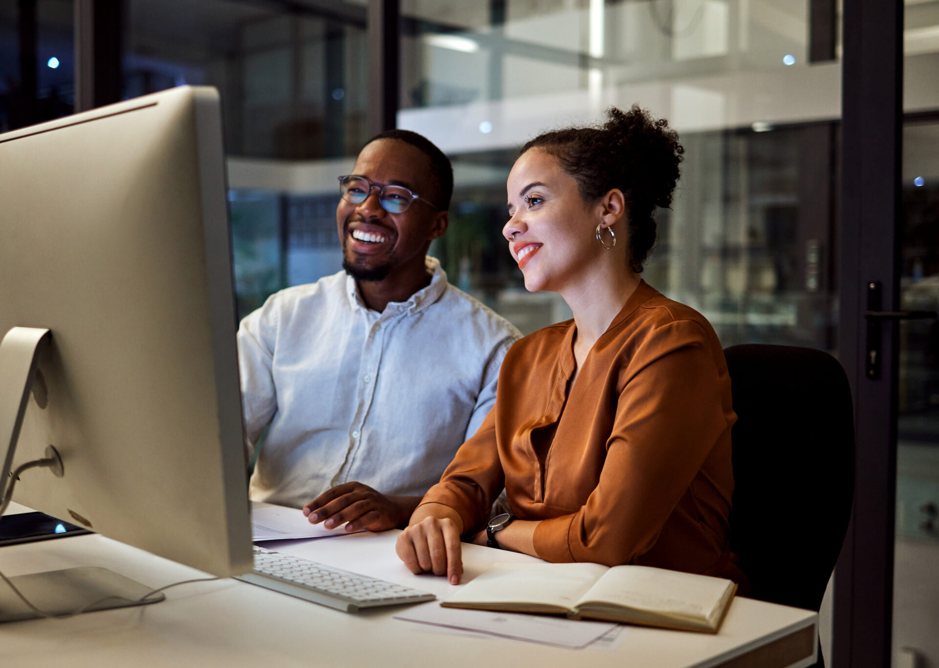 Night meeting, internet business and employees talking about web design on a computer in a dark office at night. African workers happy on company website and working overtime for partnership.