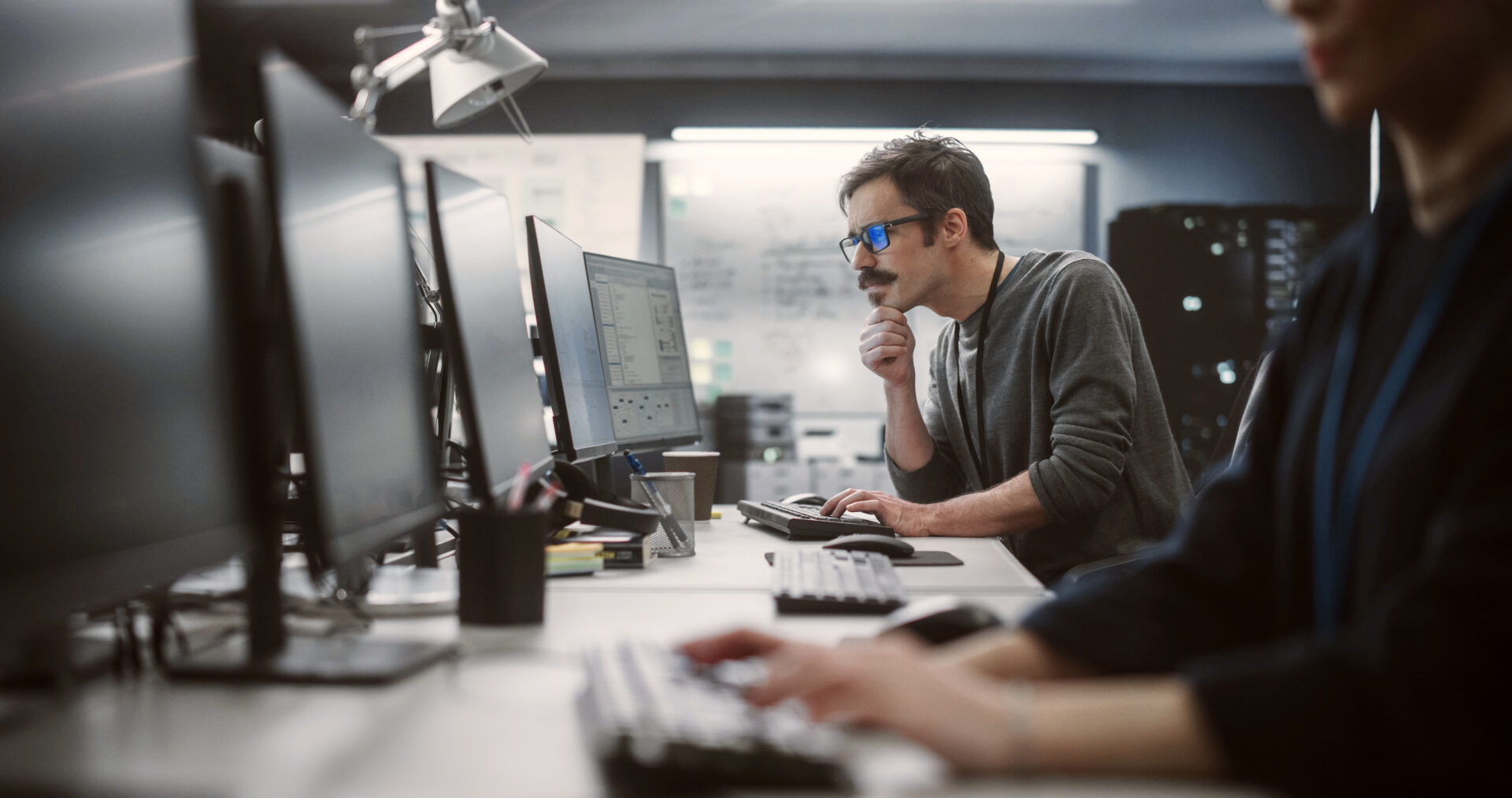 A man seated at a desk, working on two computers, focused on his tasks in a professional environment.